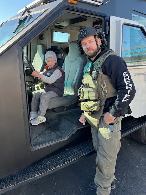 Child sitting in a truck with a member of the Sheriff's Office standing near them at the Touch a Truck Event