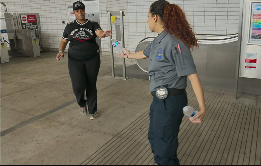 A person in a uniform offers a water bottle to another individual in a transit station.