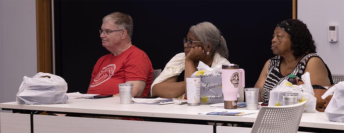 Three individuals sit at a table during what appears to be a meeting or event, with personal items and beverages in front of them.
