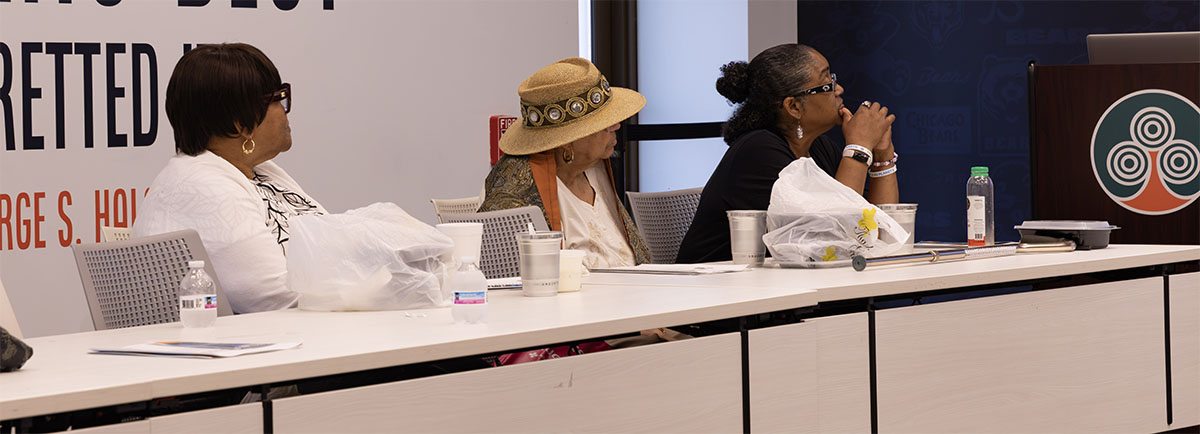 Three individuals sit at a long table during a panel discussion, each with personal items and beverages nearby.