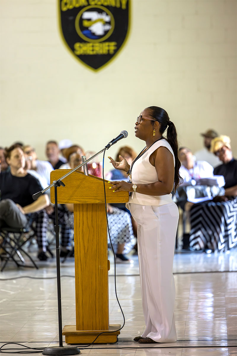 A person in a white outfit stands at a wooden podium speaking into a microphone, with an audience seated behind them and a county sheriff's badge displayed on the wall.
