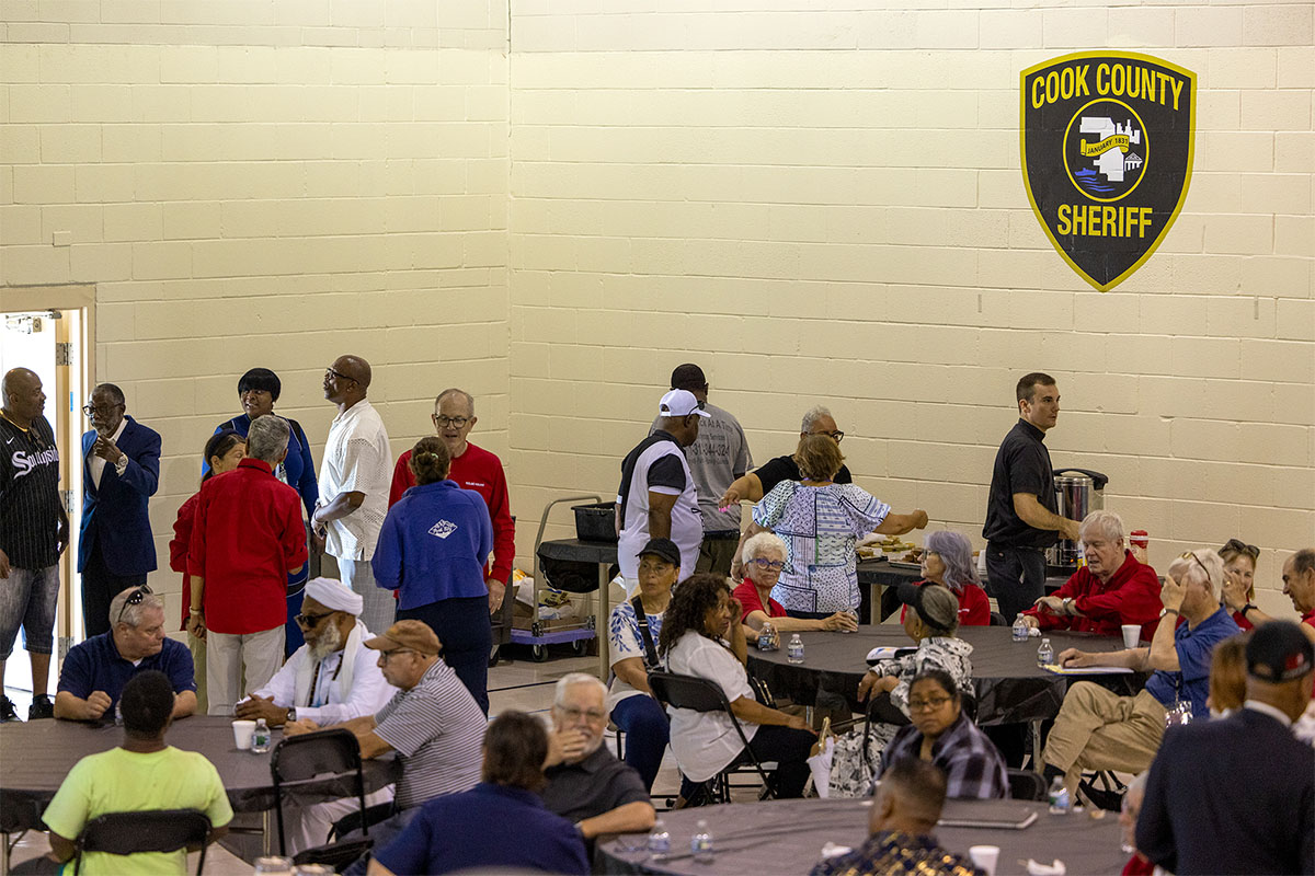 A group of people gather in a room with a Cook County Sheriff's emblem on the wall, seated at tables and standing in conversation.