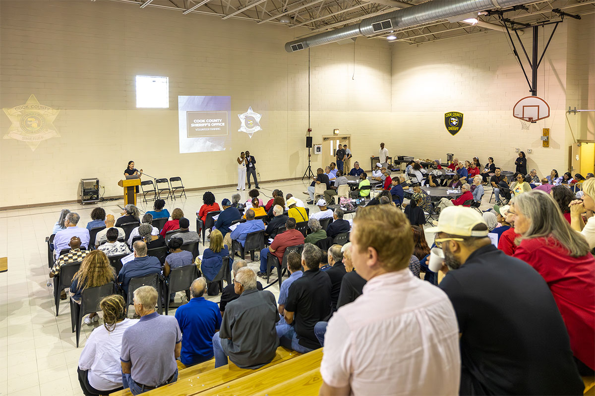 A public meeting is held in a gymnasium, with attendees seated facing a speaker at a podium under a projection screen displaying "Cook County Sheriff's Office Volunteer Program."