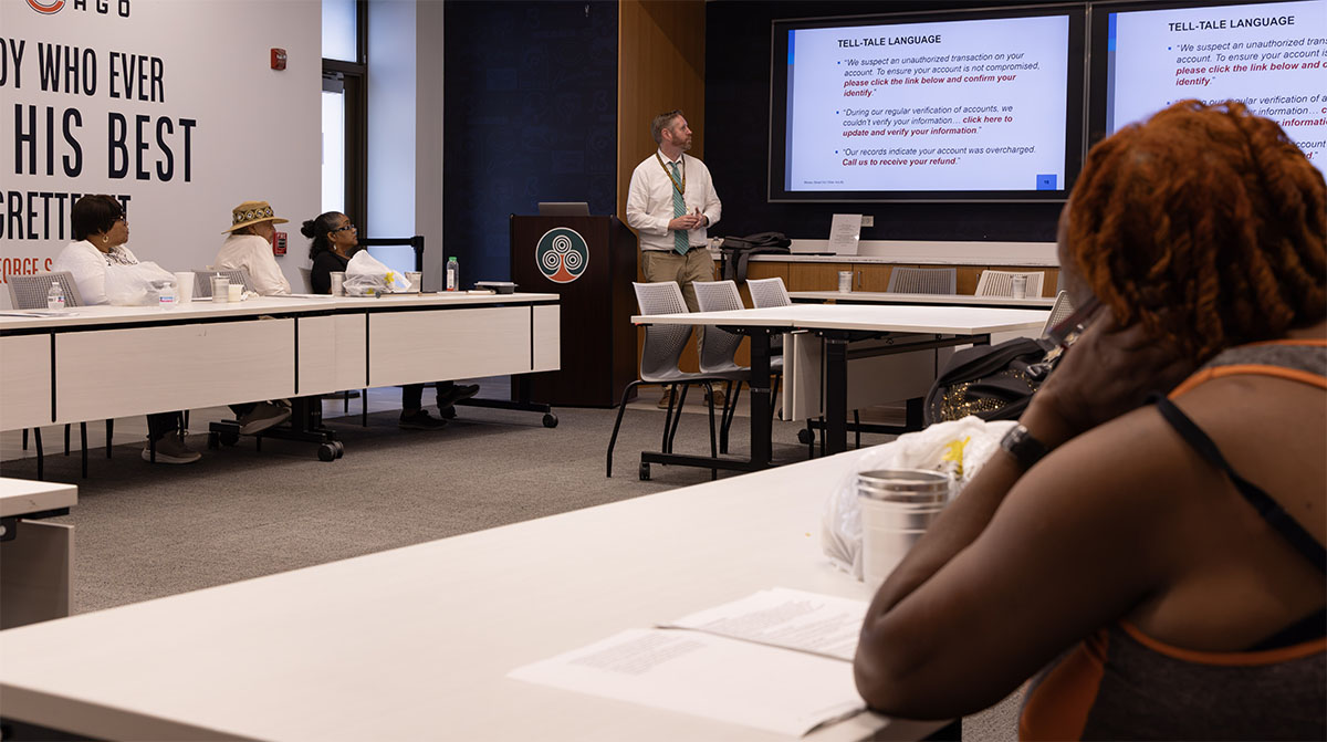 A presenter stands before an audience in a conference room, addressing attendees seated at tables while screens display text about “Tell-Tale Language” fraud alerts.