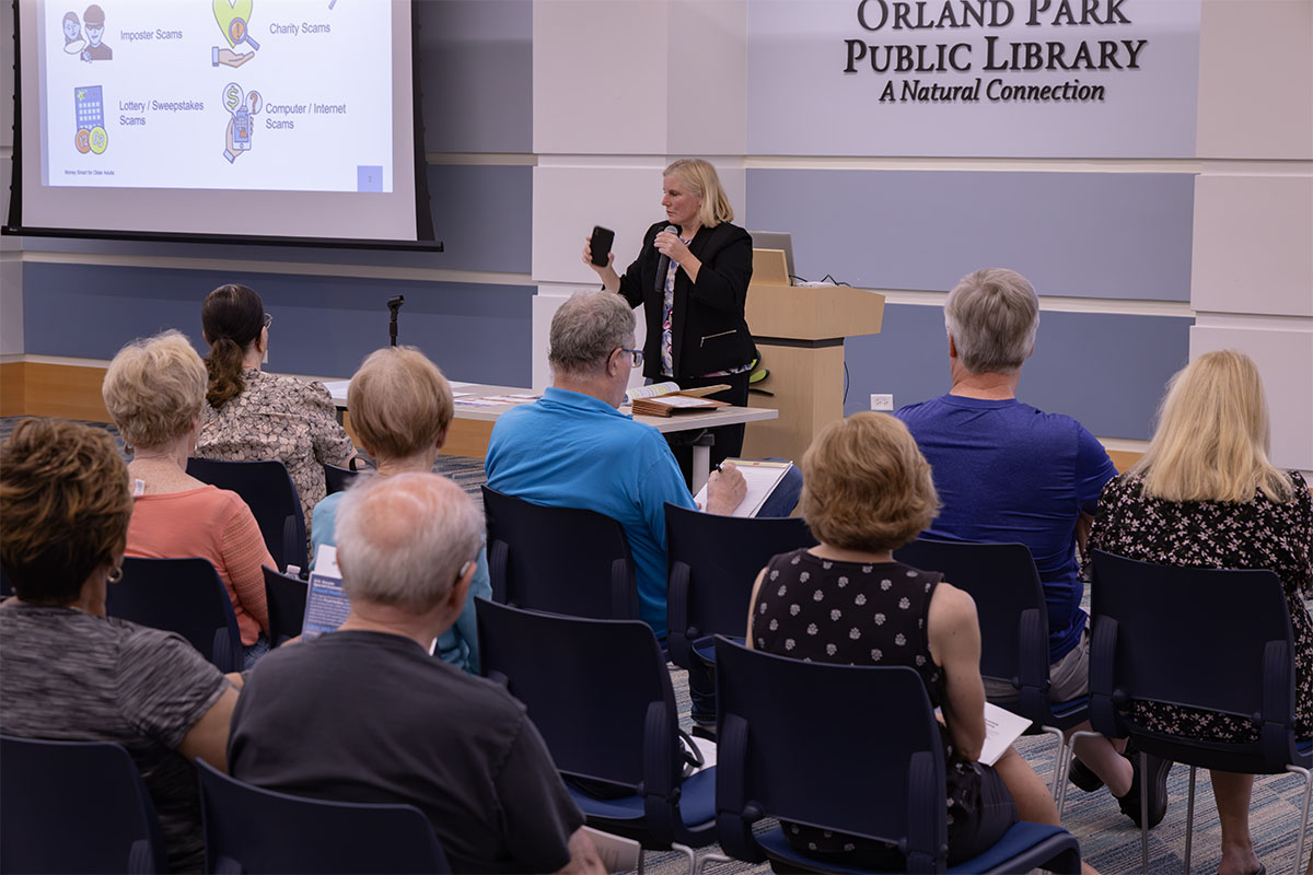 A presenter stands at a podium in front of an audience at the Orland Park Public Library, gesturing while holding a phone, with a screen displaying information about various scams.