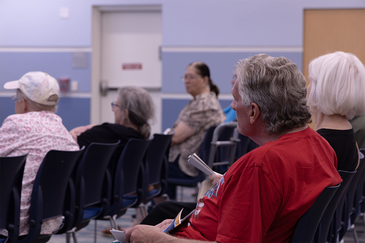 A group of people sit in rows of chairs, attentively facing forward in what appears to be a community meeting or gathering.