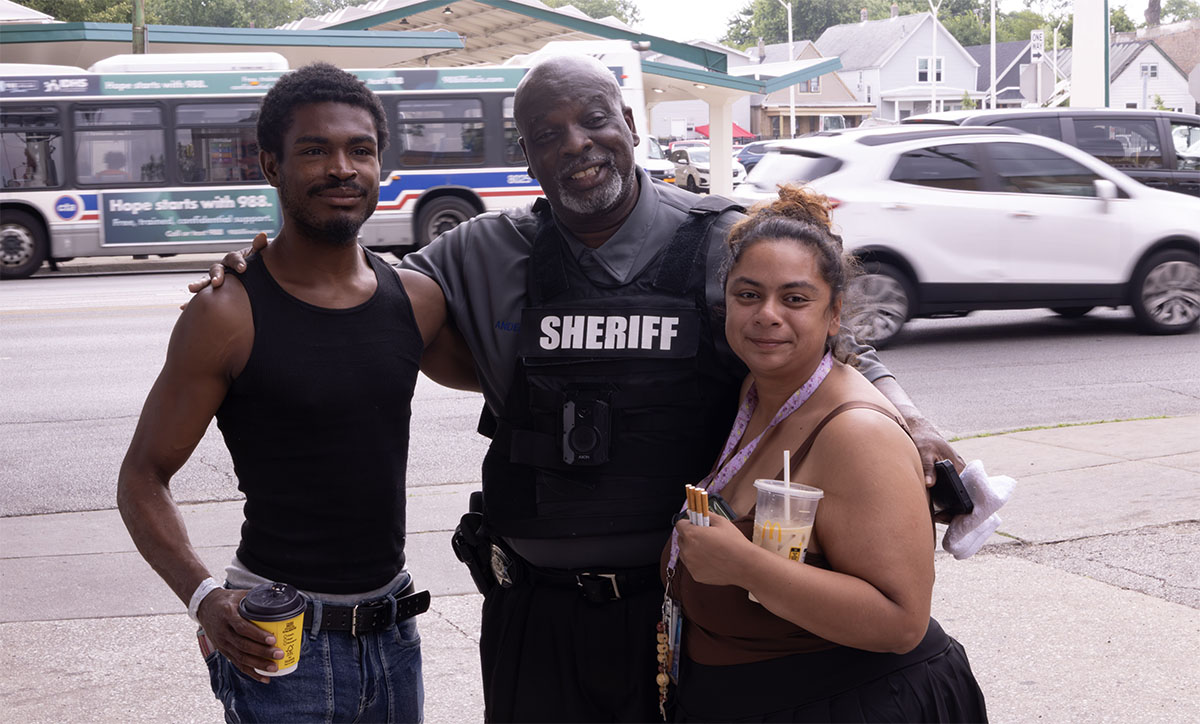 Three individuals, including a law enforcement officer, pose together on a sidewalk with drinks in hand, as vehicles pass by in the background.