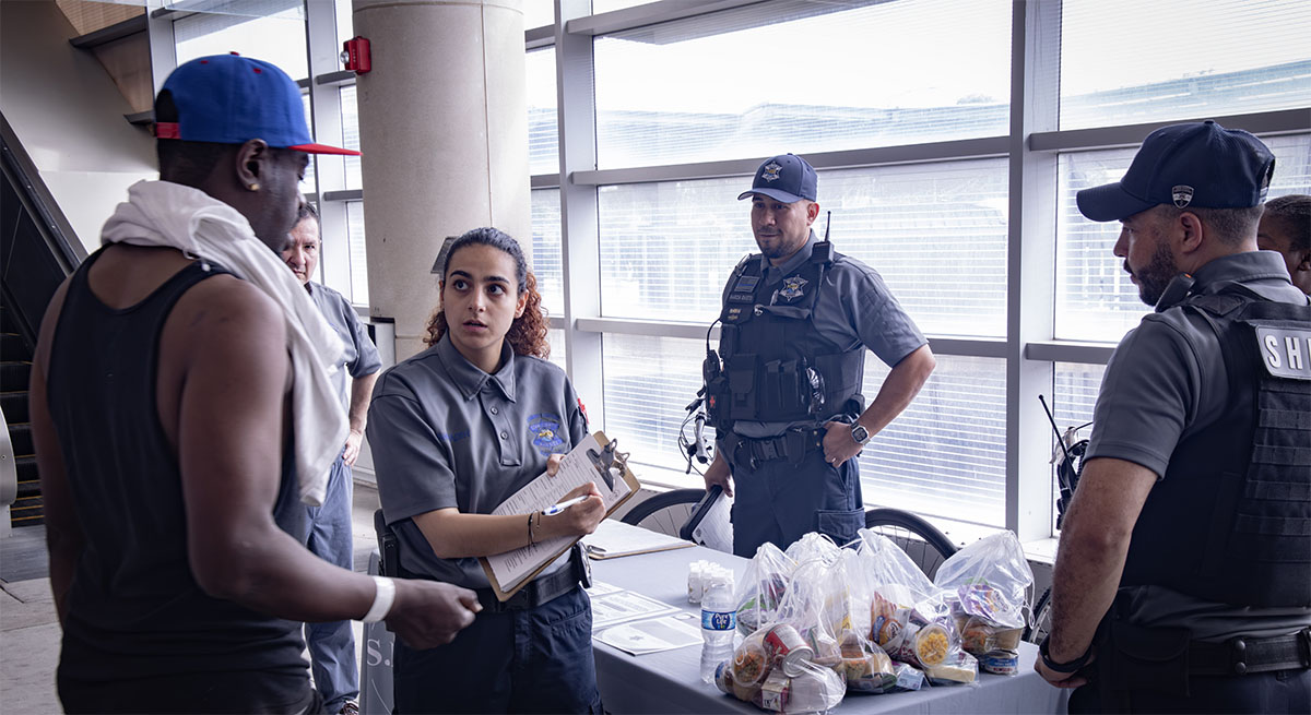 A uniformed officer interacts with a person in casual attire at a table with food items, while other officers and personnel observe nearby.