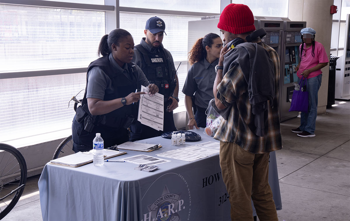 A law enforcement officer and a civilian engage in conversation at a public information booth, with other individuals nearby.