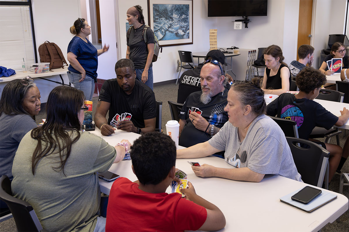 A group of people of various ages are gathered around tables playing cards in a community room.