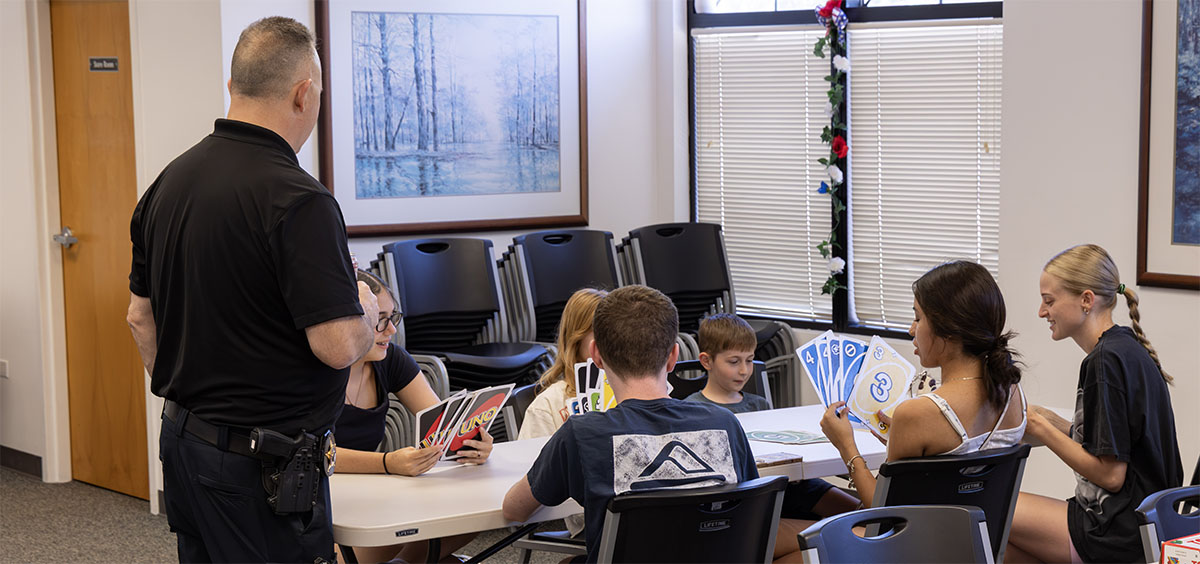 A uniformed officer stands beside a group of young people seated at a table, engaging with them as they hold and examine playing cards.