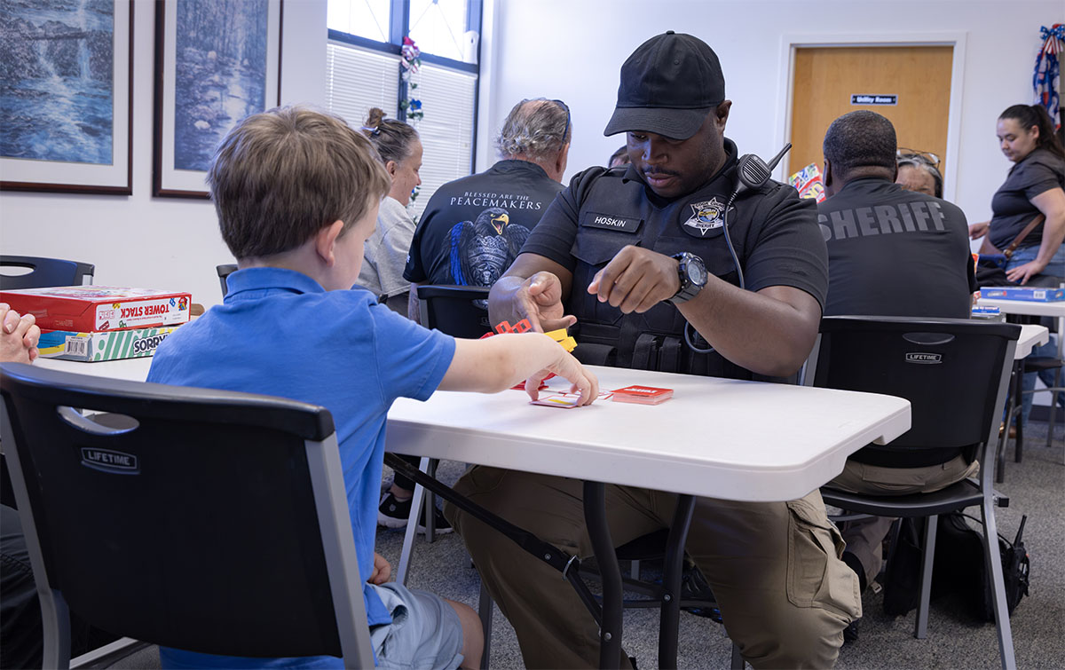 A law enforcement officer engages with a child at a table during a community event, playing a card game together.