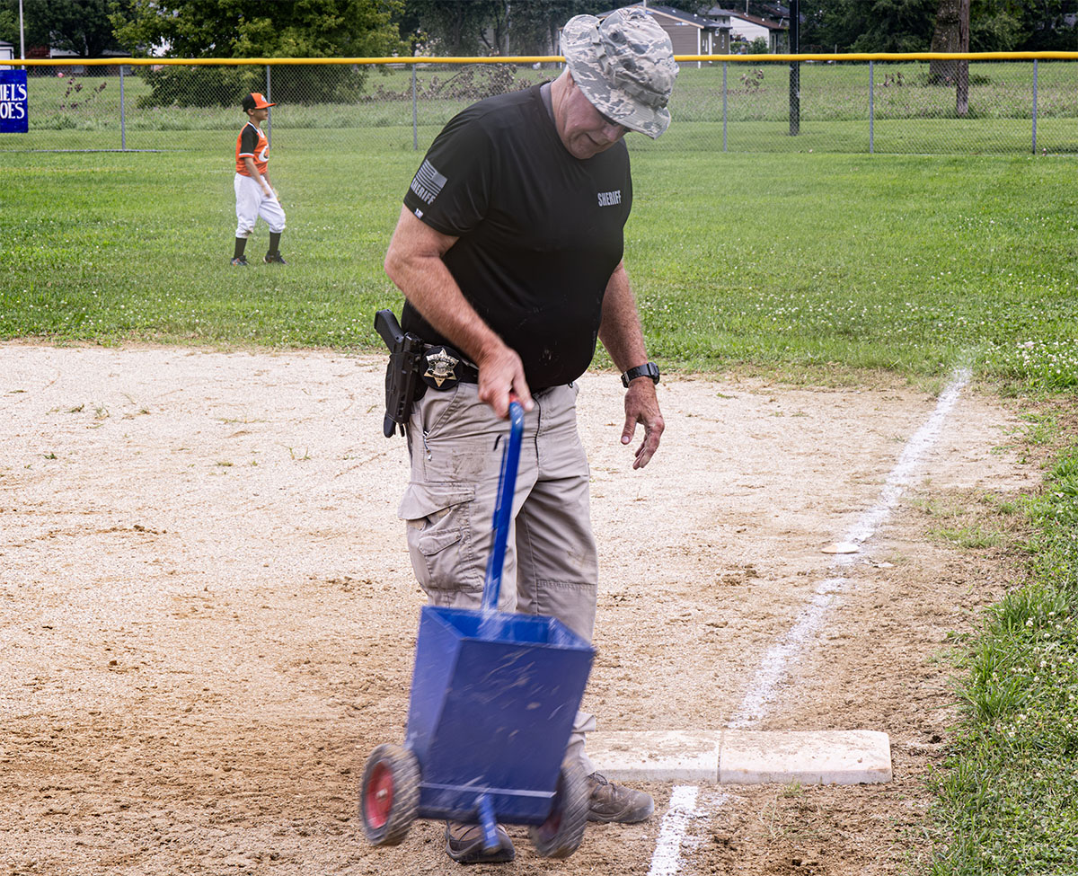 A uniformed official uses a blue marker cart to draw a white line on a baseball field's dirt area.