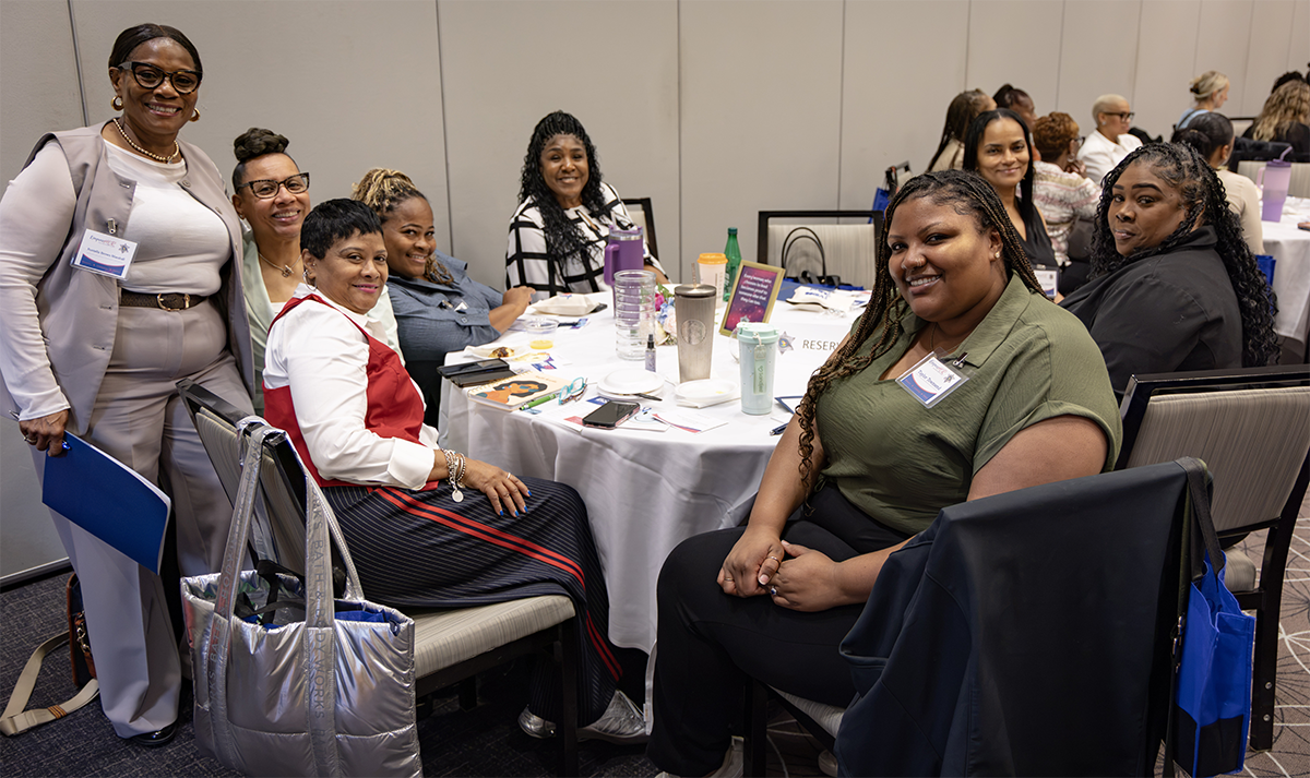 A group of individuals gather around a table at a conference or event, smiling and engaged in conversation.