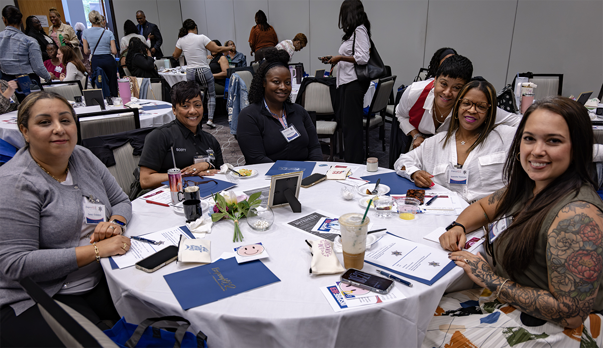 A group of individuals gather around a table at a conference or event, smiling for the camera while surrounded by materials, drinks, and personal items.