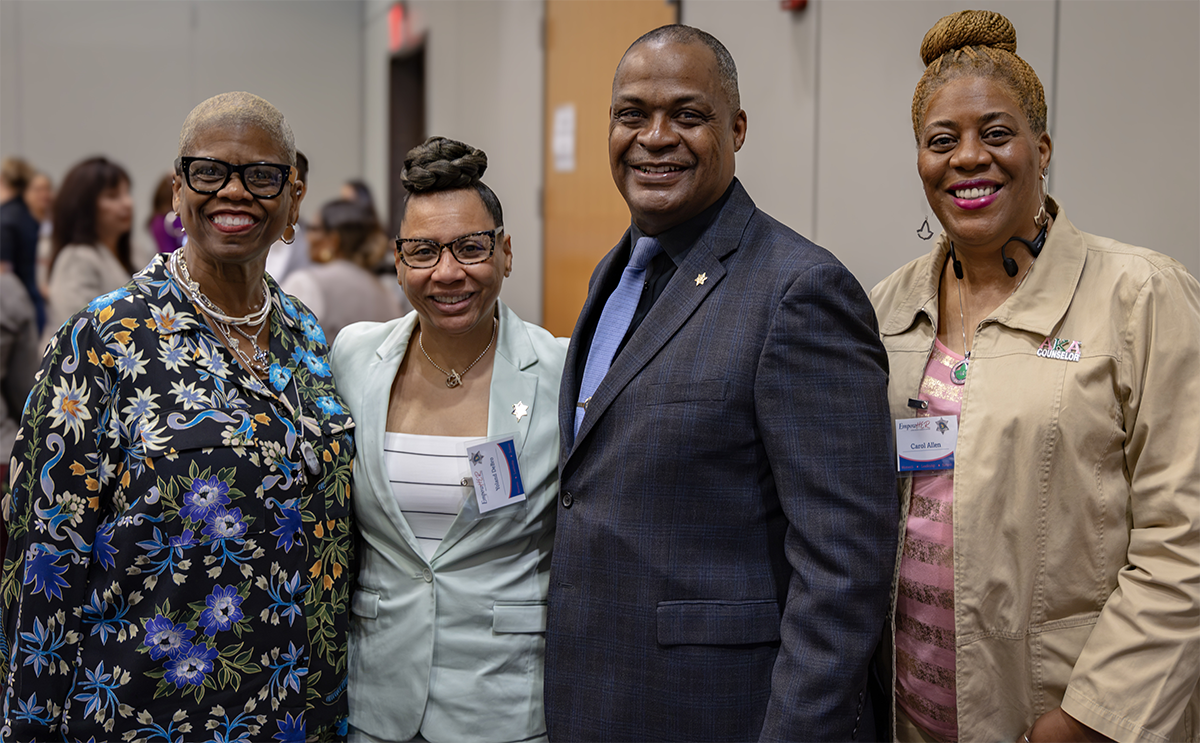 Four individuals pose together for a photo at an event, smiling and dressed in professional or semi-formal attire.