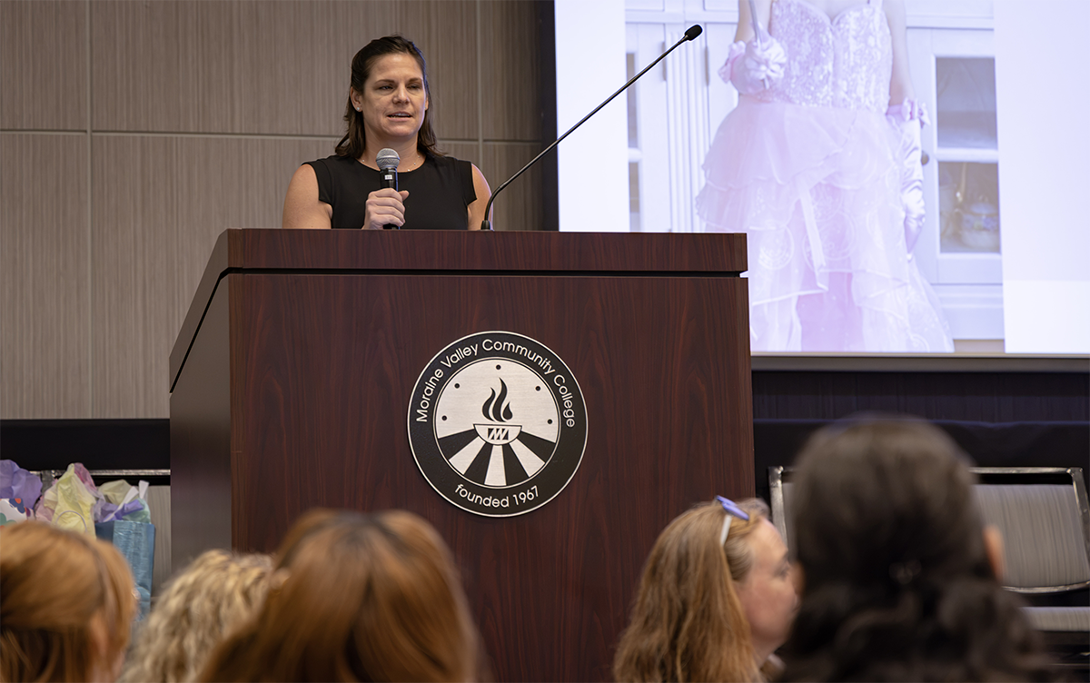 A person speaks at a podium bearing the logo of Moraine Valley Community College, with a large screen displaying a pink dress behind them.