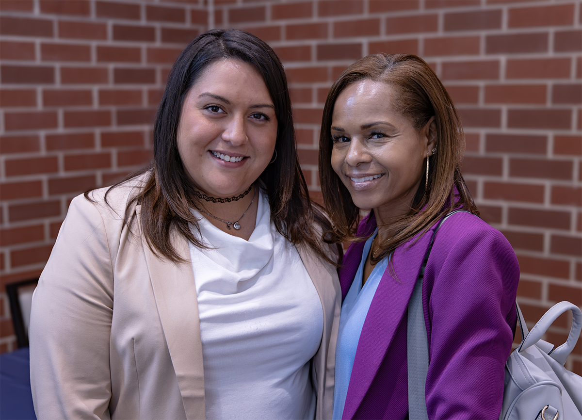Two individuals smile together in front of a brick wall, one wearing a beige blazer and the other a vibrant purple jacket.