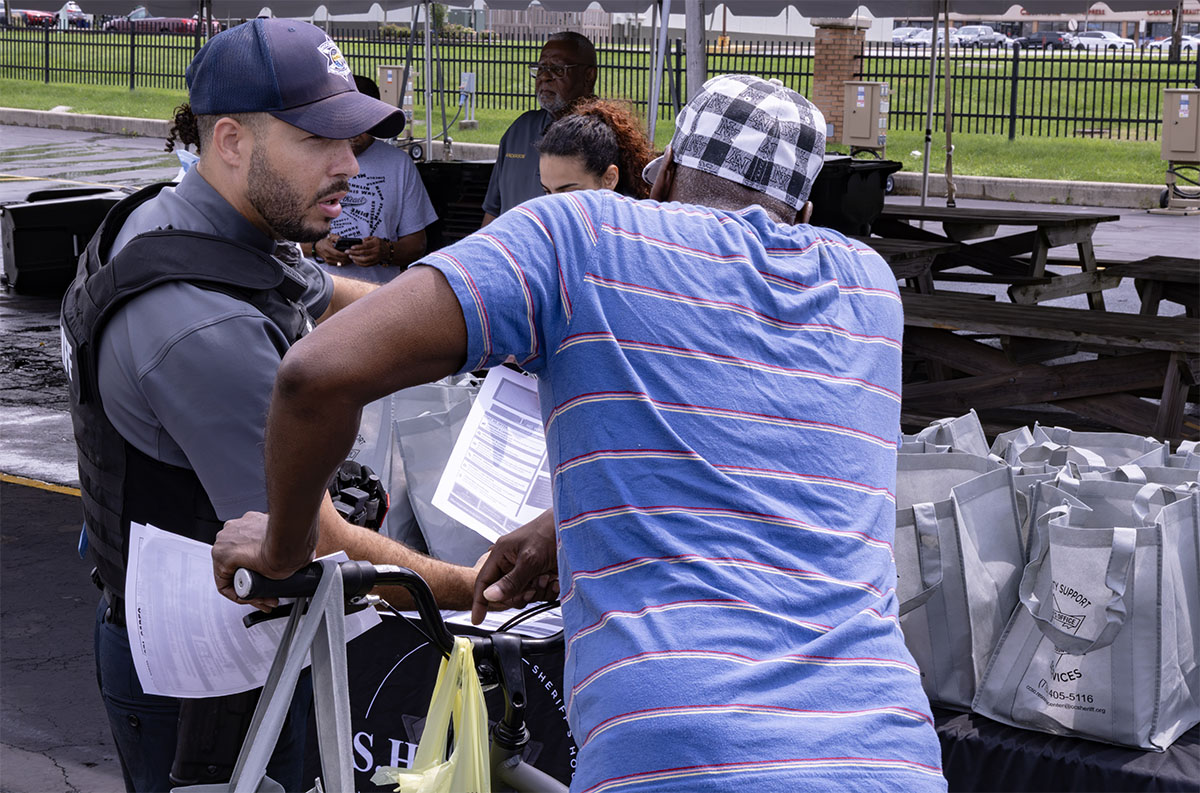 A uniformed officer interacts with a person in a striped shirt at an outdoor event, surrounded by supplies and bags.