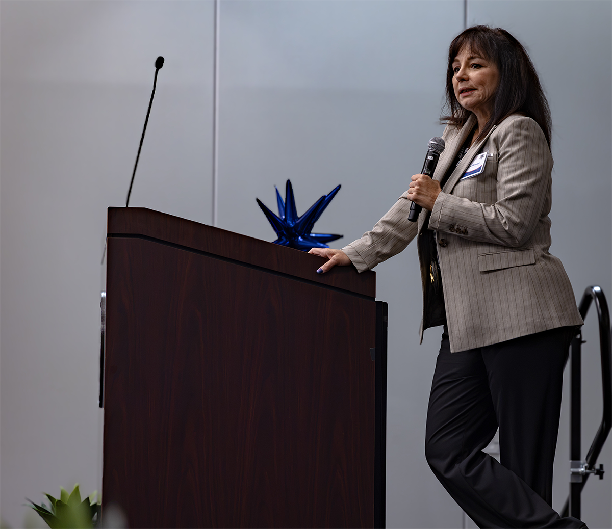 A person in a pinstripe blazer stands at a podium, holding a microphone and speaking to an audience.