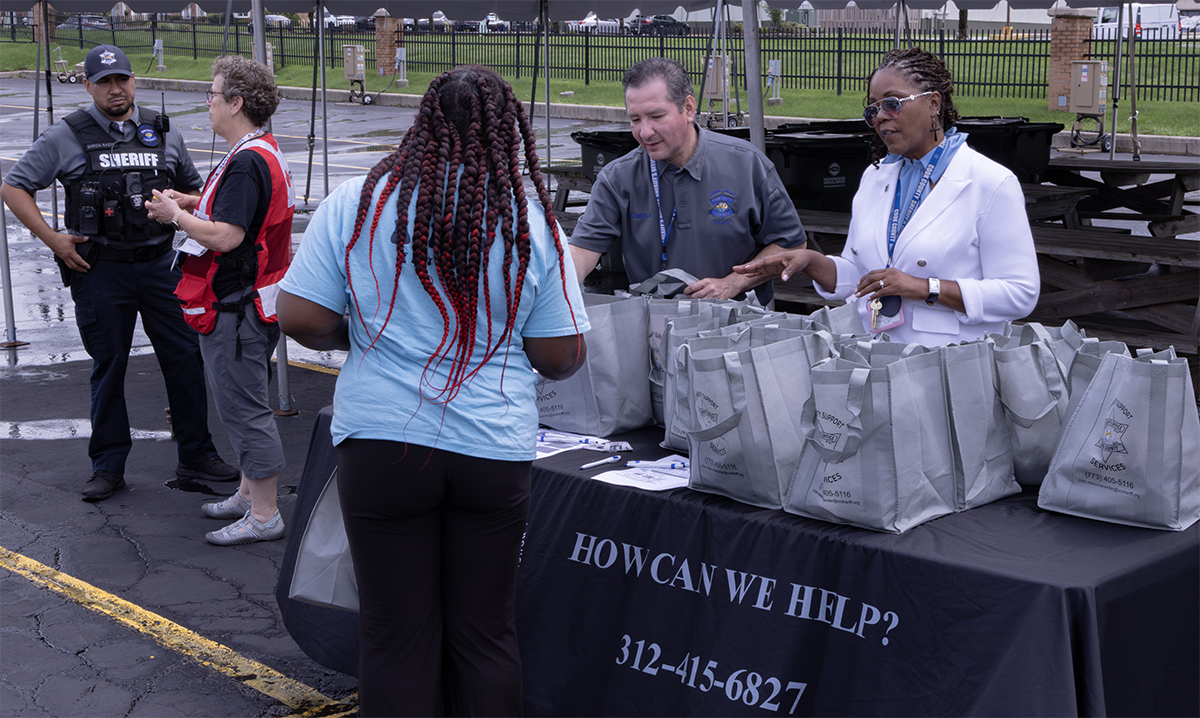 A group of people interact at an outdoor event booth offering assistance, with a table displaying bags and a sign asking "How Can We Help?" and including a phone number.