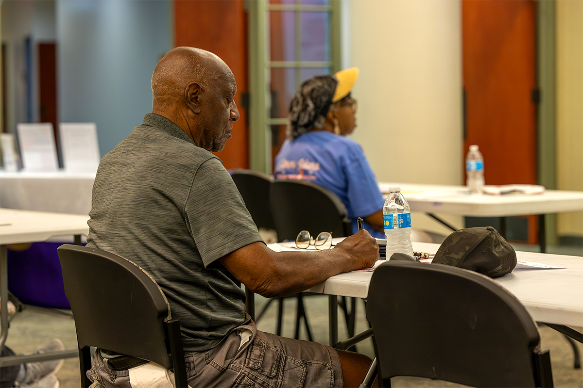 An individual sits at a table in a room, writing with a pen while wearing a gray shirt and shorts.