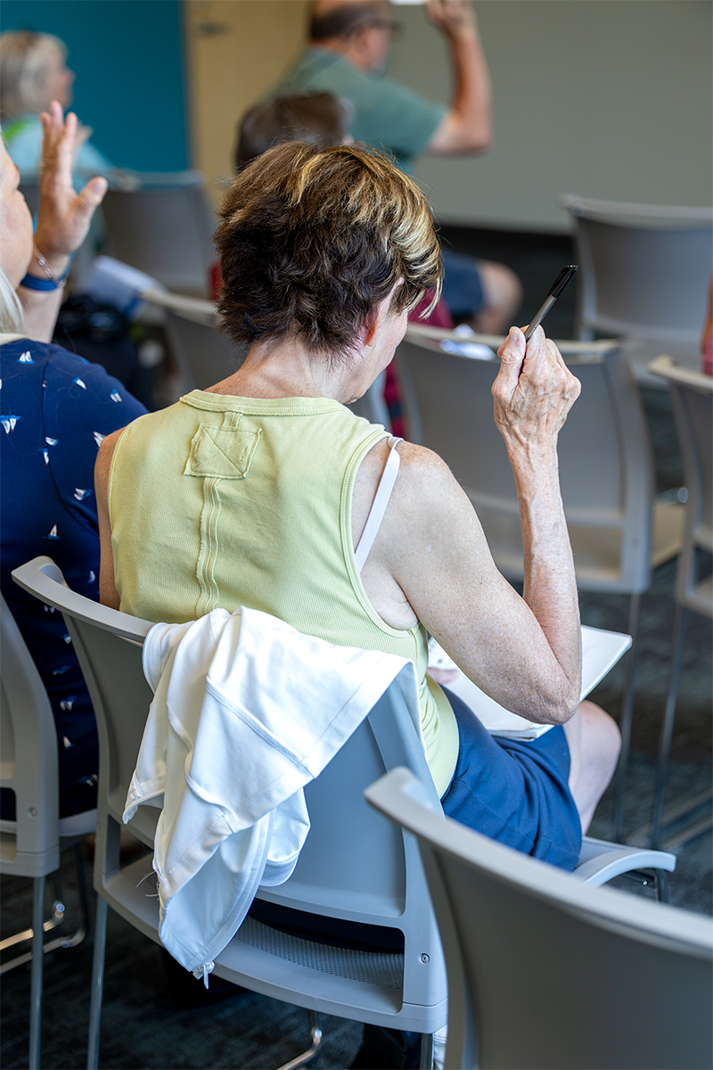 An individual with short hair sits in a classroom, holding a pen and wearing a light green sleeveless top, with a white garment draped over their chair.
